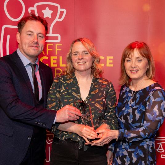 A man and two women standing in front of a large red Staff Excellence Awards banner. Lady in the middle is hold a glass trophy award.